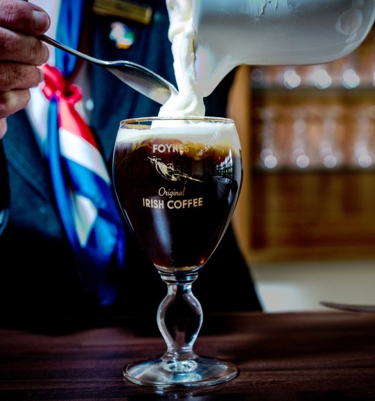 Someone pours Irish Coffee cream at Flying Boat Museum Foynes on wooden table.